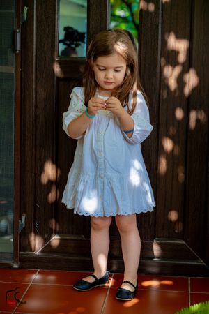 Small child in white summer dress and black shoes playing in sunlit doorway.