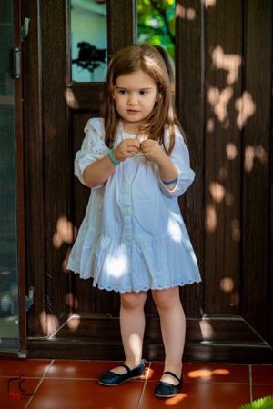 Young child in white eyelet dress standing on wooden porch with dappled sunlight.