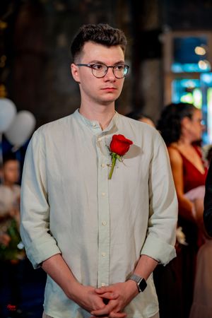 A young guest in a white button-down shirt wears glasses and a red rose boutonniere at a formal event.