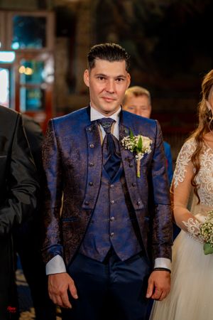 A groom wearing an elegant blue patterned suit with matching vest and tie stands at a formal wedding ceremony.