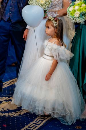 A flower girl in a white tulle dress stands with a balloon during a wedding ceremony.