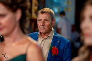 Formal event attendee in blue blazer and tan shirt with red boutonniere in decorated venue.