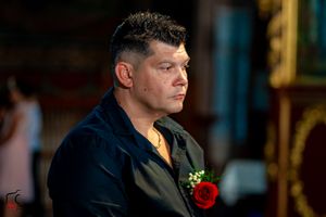 Guest at formal event wearing dark suit with red boutonniere in ornate church setting.