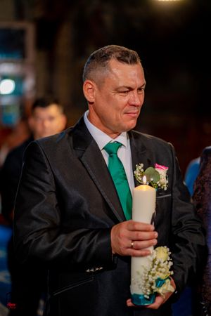 Person in black suit with green tie holding a decorated candle during ceremony.