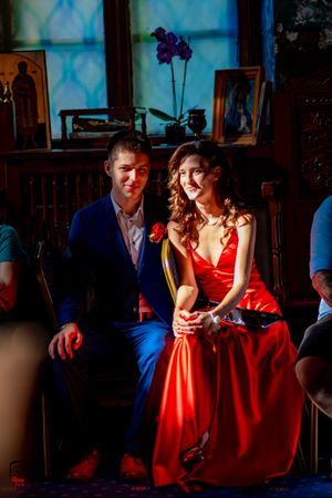 Couple in formal attire sits together with dramatic lighting and purple orchid in background.