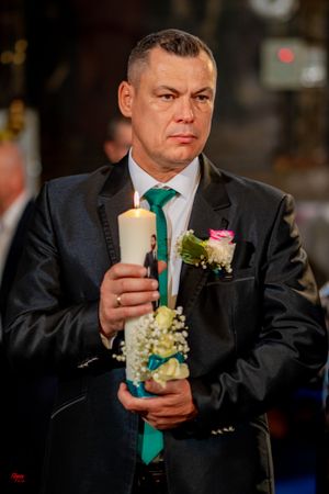Person in dark suit and green tie holds decorated ceremonial candle during formal event.