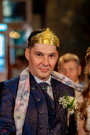 Groom wearing navy patterned suit and golden crown during traditional ceremony.