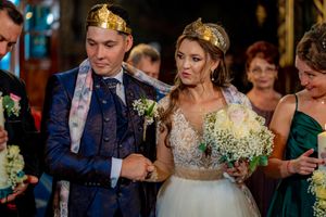 Couple wearing golden crowns during wedding ceremony with guests holding candles in ornate venue.