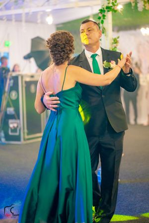 Formal dance partners in elegant attire performing under decorative lighting and greenery.