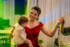 Woman in red dress dancing while holding a small child at an indoor celebration.