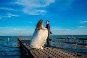 Romantic waterfront scene with wedding couple on wooden dock against bright blue horizon.
