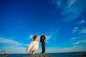 A couple in wedding attire stands against a vibrant blue sky on a wooden pier by the ocean.