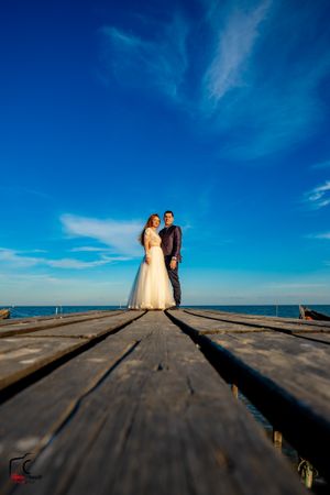 Wedding couple stands at end of wooden pier with ocean and blue sky backdrop.