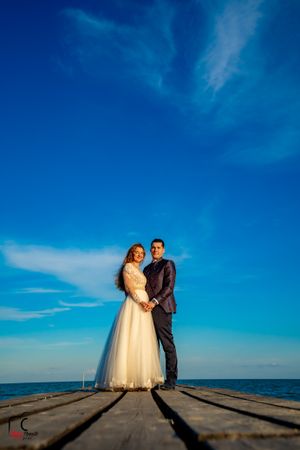 Wedding couple poses on wooden dock with dramatic shadows and ocean backdrop.