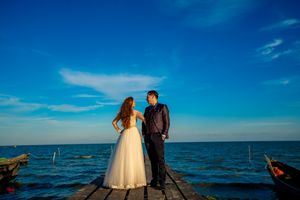 Wedding couple embraces on seaside pier with bright blue sky and ocean in background.