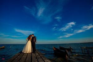 Silhouetted figures in wedding attire stand on a wooden pier overlooking the ocean at sunset.