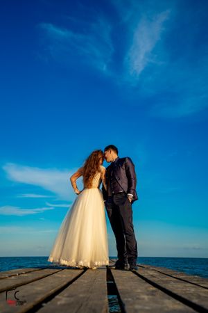 Wedding couple poses on wooden dock with dramatic blue sky and ocean horizon.