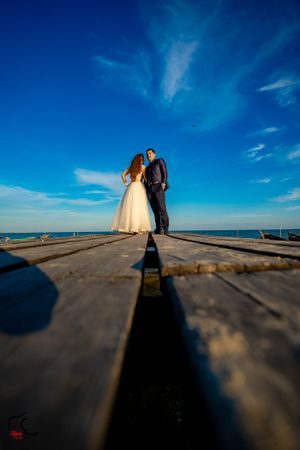 A couple in wedding attire stands on a wooden pier against a vibrant blue sky and ocean backdrop.