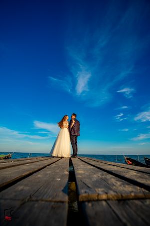 A couple standing on weathered wooden planks with the ocean and bright blue sky as backdrop.