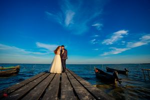 A couple standing on a wooden pier overlooking the ocean with boats floating nearby at sunset.
