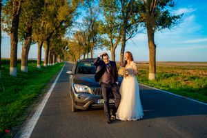 A wedding couple stands beside their car on a beautiful tree-lined rural road.
