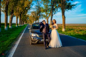 A couple posing with their car on a scenic tree-lined road during a wedding photoshoot.