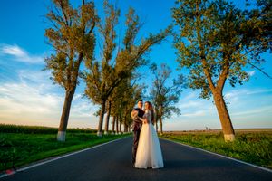 A couple in wedding attire stands on a tree-lined country road against a bright blue sky at sunset.
