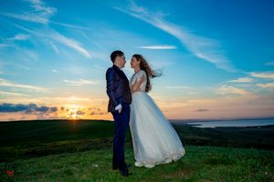 Wedding couple shares intimate moment on hilltop with wind-swept dress and scenic backdrop.