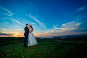A couple in wedding attire poses on a hilltop meadow against a dramatic sunset sky.