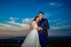 Newlyweds share romantic moment on scenic hilltop with dramatic sunset clouds behind them.