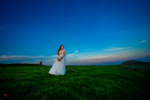 A couple in wedding attire poses on a hilltop against a vibrant blue sky and green landscape at sunset.