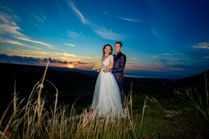 Newlyweds pose on grassy hillside with dramatic blue sky and clouds in background.