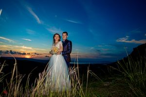 Romantic sunset wedding portrait with couple standing on hilltop overlooking coastal landscape.
