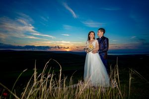 Couple in wedding attire poses on mountainside with dramatic sunset colors.