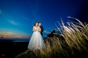 Couple in wedding attire poses against dramatic sunset sky and mountain landscape.