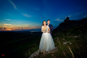 Couple in wedding attire stands on rocky overlook during beautiful sunset.