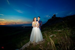 Wedding couple poses on mountain peak during dramatic sunset with castle silhouette.