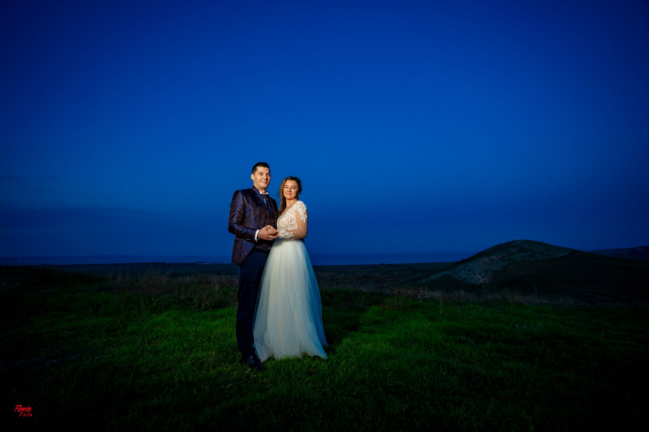 A wedding couple stands on a grassy hilltop against a deep blue twilight sky.
