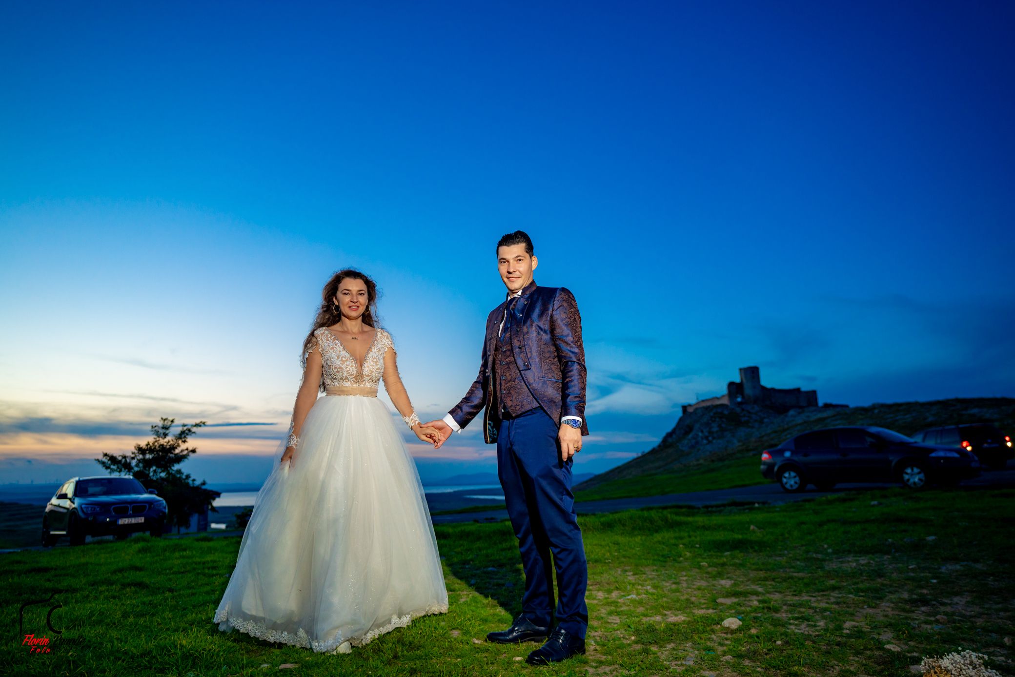 Wedding couple poses against dramatic blue sky in outdoor countryside setting at dusk.