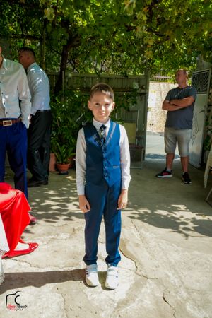 Person in blue vest and formal attire standing outdoors under leafy pergola.