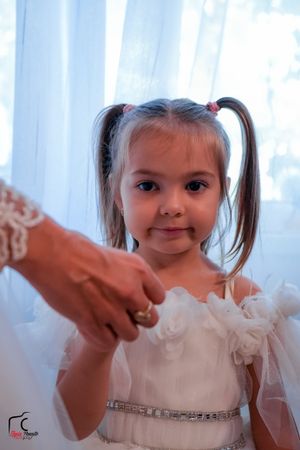 A young child in white dress making playful expressions against light curtains.