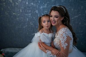 Sweet moment between a bride in lace gown and flower girl in white dress against blue backdrop.