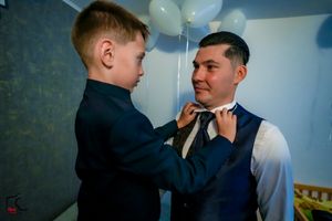 A touching pre-event moment showing bow tie adjustment against a blue wall with balloons.