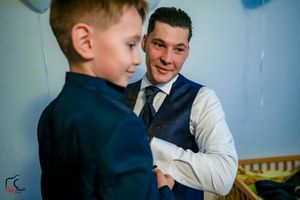 A heartwarming interaction between an adult in formal wear and a child in a navy suit against a blue wall with balloons.