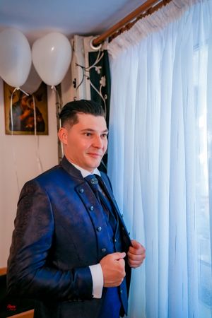 Person in formal attire stands near white curtains and balloons.