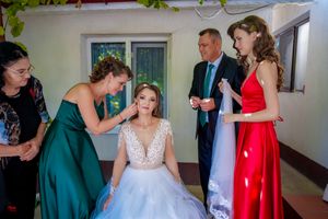 Wedding party helps prepare the bride in a sunlit room with green foliage visible through windows.