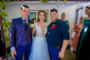 Wedding party members pose together in formal attire against a white wall with greenery.