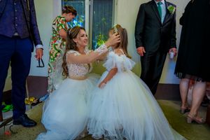 Two people in flowing white dresses share a playful dance moment at a celebration.