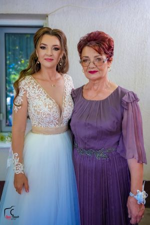 Two women embrace while wearing formal dresses - one in white lace and tulle, the other in elegant purple.