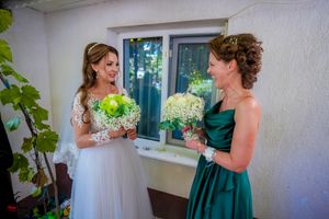 A bride in a white lace gown and bridesmaid in emerald green dress hold matching floral bouquets.